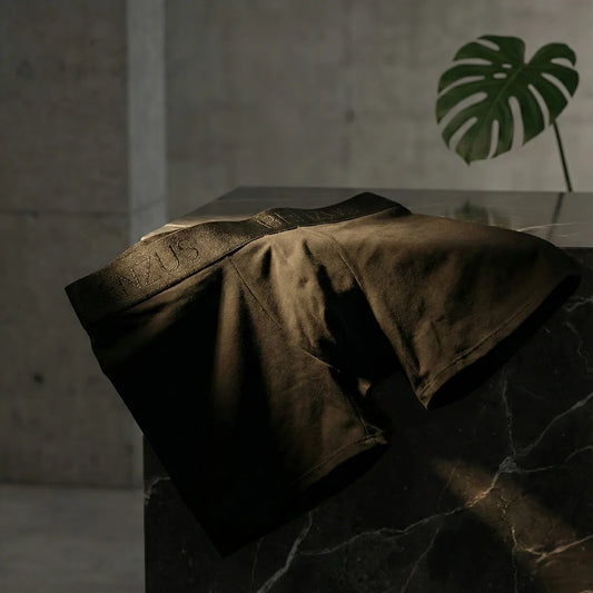 Fotografía estética dark de lujo: Bóxer negro BENZUS ORIGEN sobre superficie de mármol oscuro con iluminación dramática. Se destaca la textura suave del algodón Pima, el diseño elegante del elástico tono sobre tono y el ambiente sofisticado.