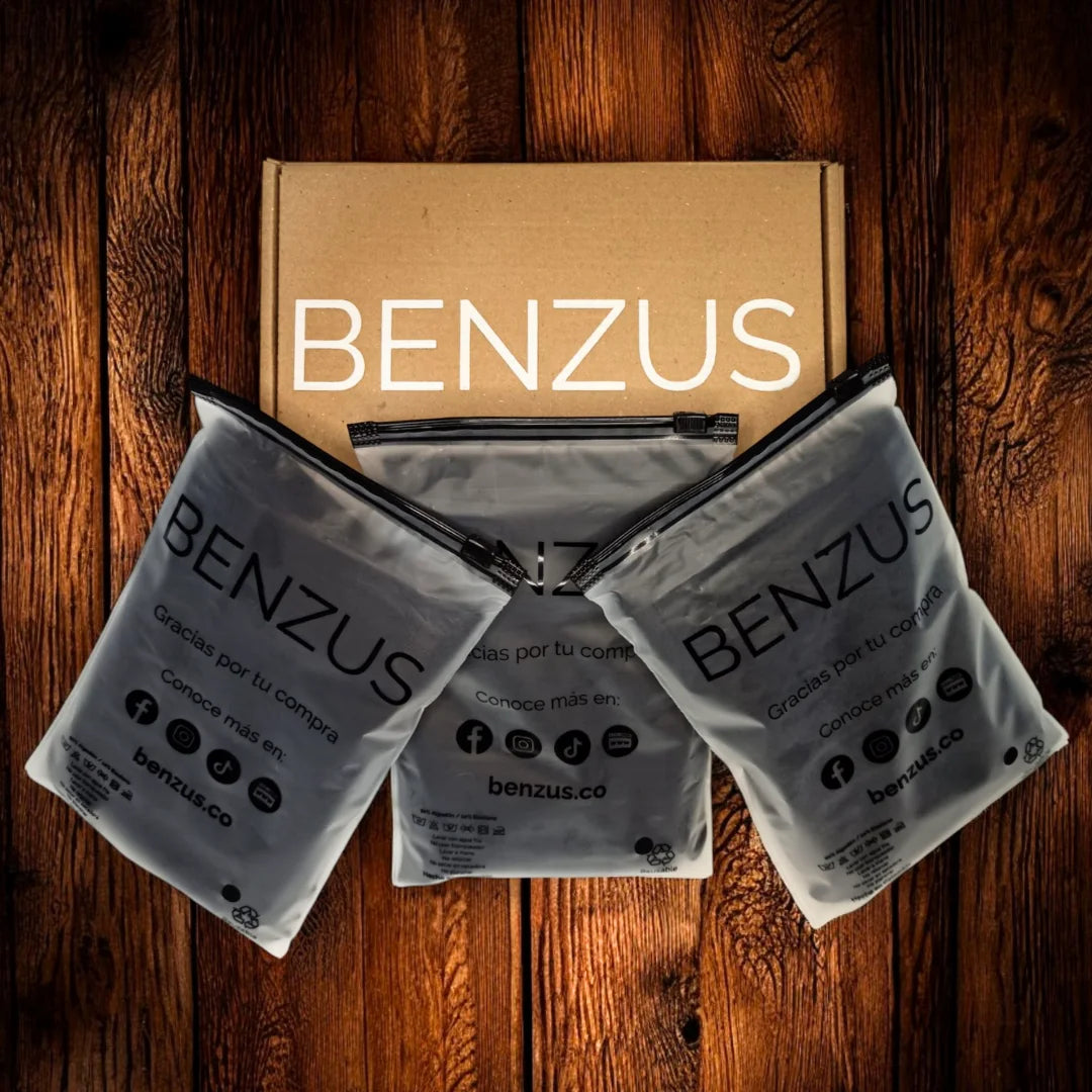 Vista cenital del empaque premium del Tri-Pack BENZUS sobre superficie de madera. Muestra la caja de envío ecológica con el logo en blanco y tres bolsas individuales de seguridad con acabado mate esmerilado y cierre hermético, que contienen los boxers. Detalles visibles de agradecimiento y redes sociales en el empaque de las 3 bolsas.